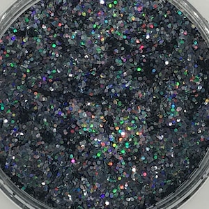 Op de afbeelding: Een doorzichtige plastic container gevuld met donkergrijze glitter. De glitterstukjes zijn zeshoekig en reflecteren meerdere kleuren. De container staat op een witte ondergrond.