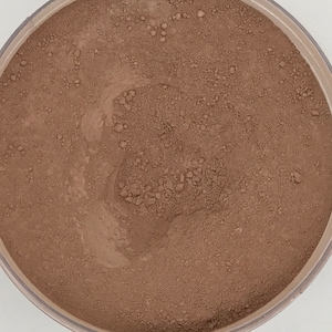 May include: A close-up view of a brown powder in a round container. The powder is finely ground and has a smooth texture.