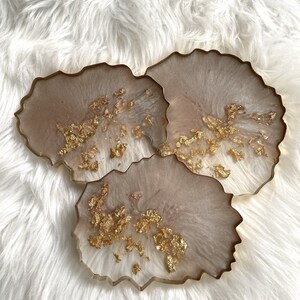 May include: Set of three beige resin coasters with gold leaf accents. The coasters have an irregular, organic shape.