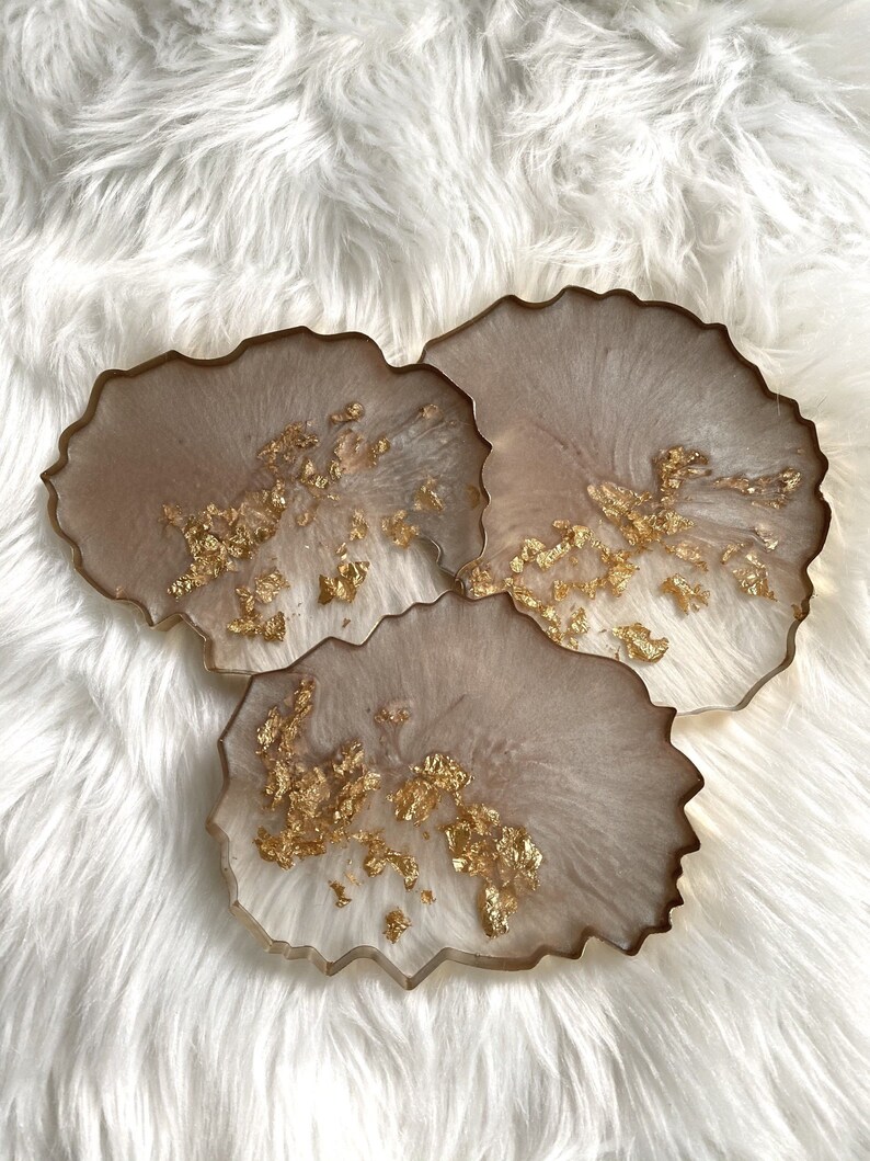 May include: Set of three beige resin coasters with gold leaf accents. The coasters have an irregular, organic shape.