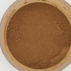 May include: A close-up view of a brown powder in a clear plastic container. The powder has a slightly grainy texture and is evenly distributed throughout the container.