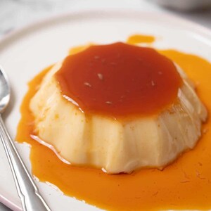 Può includere: Primo piano di un flan cremoso ricoperto da una ricca salsa al caramello su un piatto bianco con un cucchiaio d'argento.