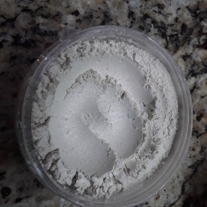 Puede incluir: Primer plano de un polvo blanco en un recipiente de plástico transparente. El polvo está finamente molido y tiene una textura suave.