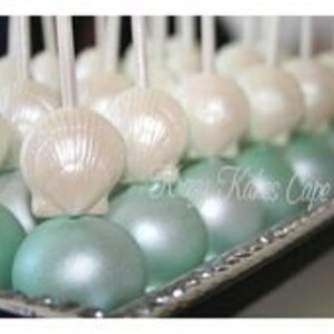 K&ouml;nnte beinhalten: Ein Tablett mit wei&szlig;en und t&uuml;rkisfarbenen Cake Pops, die mit perlmuttartigen Muscheln verziert sind. Die Cake Pops sind in Reihen auf einem silbernen Tablett angeordnet.
