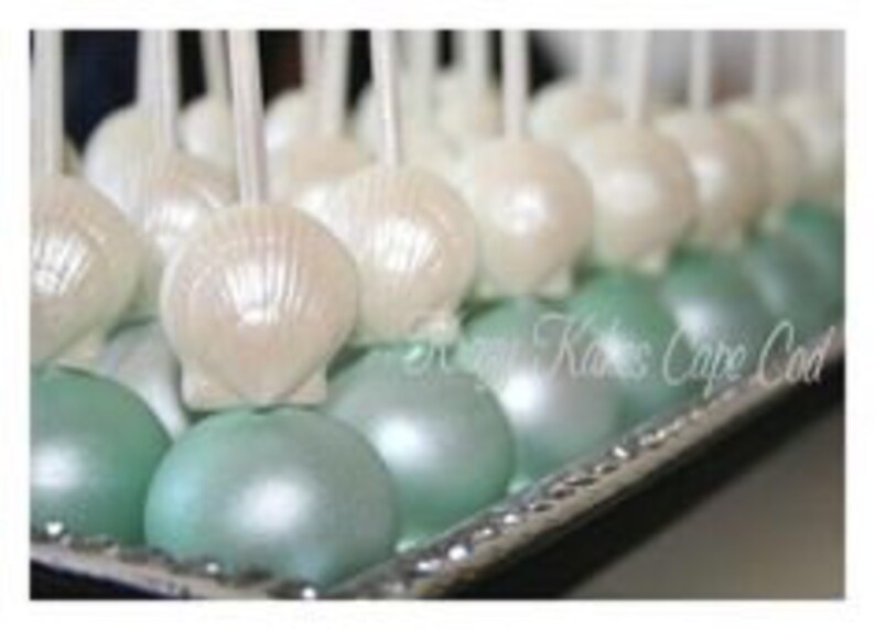 K&ouml;nnte beinhalten: Ein Tablett mit wei&szlig;en und t&uuml;rkisfarbenen Cake Pops, die mit perlmuttartigen Muscheln verziert sind. Die Cake Pops sind in Reihen auf einem silbernen Tablett angeordnet.