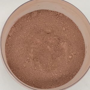 May include: A close-up of a brown powder in a white plastic container. The powder is finely ground and has a smooth texture.