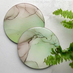 May include: Two round coasters featuring an abstract design in shades of green and brown. The coasters have a glossy finish and are made with resin.