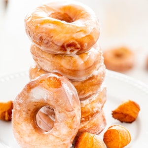 Pode incluir: Uma pilha de seis donuts glaciados num prato branco. Os donuts estão dispostos em forma de torre, com o donut maior na parte inferior e o donut menor na parte superior. Há também várias pequenas massas redondas de cor laranja no prato.