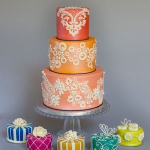 Peut inclure: Un g&acirc;teau de mariage &agrave; trois &eacute;tages d&eacute;cor&eacute; de dentelle blanche et d'accents de perles. Le g&acirc;teau est un d&eacute;grad&eacute; d'orange et de rose. Le g&acirc;teau est entour&eacute; de six petits g&acirc;teaux, chacun d&eacute;cor&eacute; de couleurs et de motifs diff&eacute;rents.