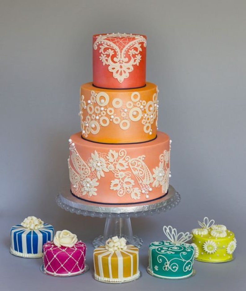 Peut inclure: Un g&acirc;teau de mariage &agrave; trois &eacute;tages d&eacute;cor&eacute; de dentelle blanche et d'accents de perles. Le g&acirc;teau est un d&eacute;grad&eacute; d'orange et de rose. Le g&acirc;teau est entour&eacute; de six petits g&acirc;teaux, chacun d&eacute;cor&eacute; de couleurs et de motifs diff&eacute;rents.