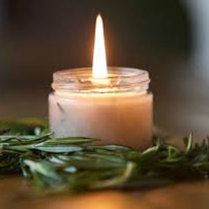 May include: A small, clear glass jar candle with white wax and a single flame burning. The candle is surrounded by green rosemary sprigs.