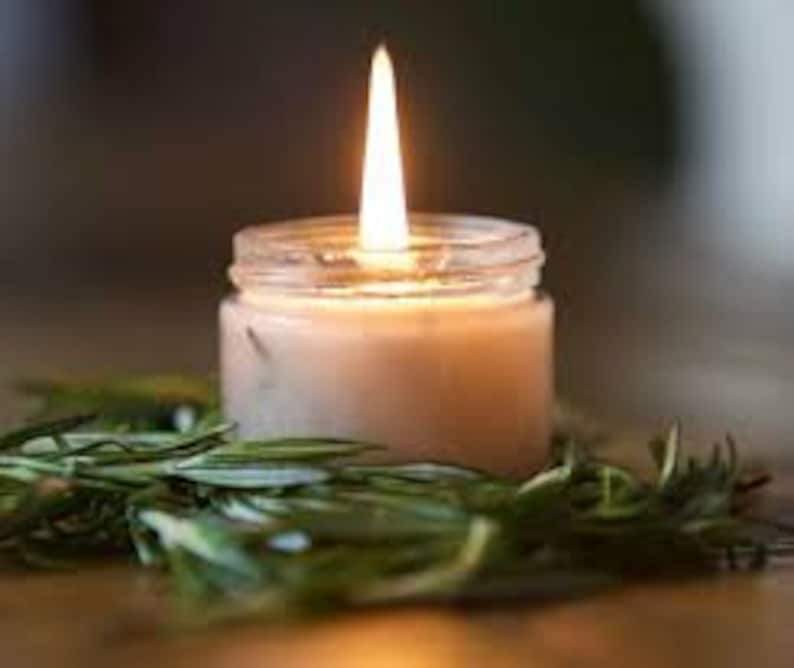 May include: A small, clear glass jar candle with white wax and a single flame burning. The candle is surrounded by green rosemary sprigs.