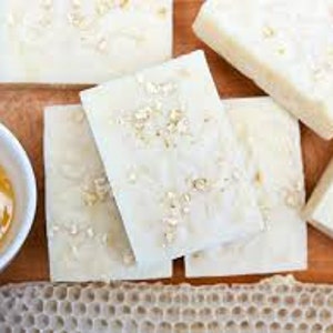 May include: Four bars of white soap with oatmeal and honey. The soap is on a wooden surface with a bowl of honey and a piece of honeycomb.