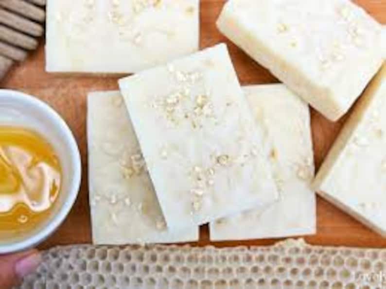 May include: Four bars of white soap with oatmeal and honey. The soap is on a wooden surface with a bowl of honey and a piece of honeycomb.