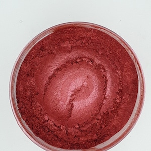 Puede incluir: Un pequeño recipiente de polvo de mica rojo. El polvo es fino y tiene un ligero brillo.