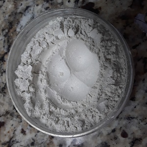 Puede incluir: Primer plano de un polvo blanco en un frasco de vidrio transparente. El polvo es de color gris claro y tiene una textura suave.