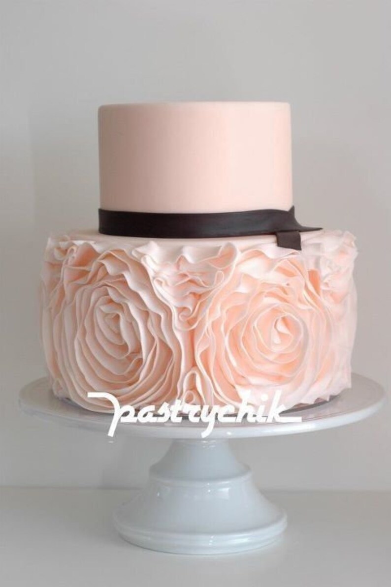 Peut inclure: Un g&acirc;teau de mariage &agrave; deux &eacute;tages avec un gla&ccedil;age rose p&acirc;le et un ruban noir. L'&eacute;tage inf&eacute;rieur est d&eacute;cor&eacute; d'un motif pliss&eacute;.