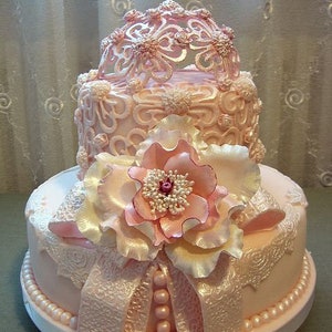 Peut inclure: Un g&acirc;teau de mariage &agrave; deux &eacute;tages de couleur rose, d&eacute;cor&eacute; de dentelle et de perles. Le niveau sup&eacute;rieur pr&eacute;sente un d&ocirc;me ajour&eacute; d&eacute;licat, tandis que le niveau inf&eacute;rieur est orn&eacute; d'une grande fleur rose.