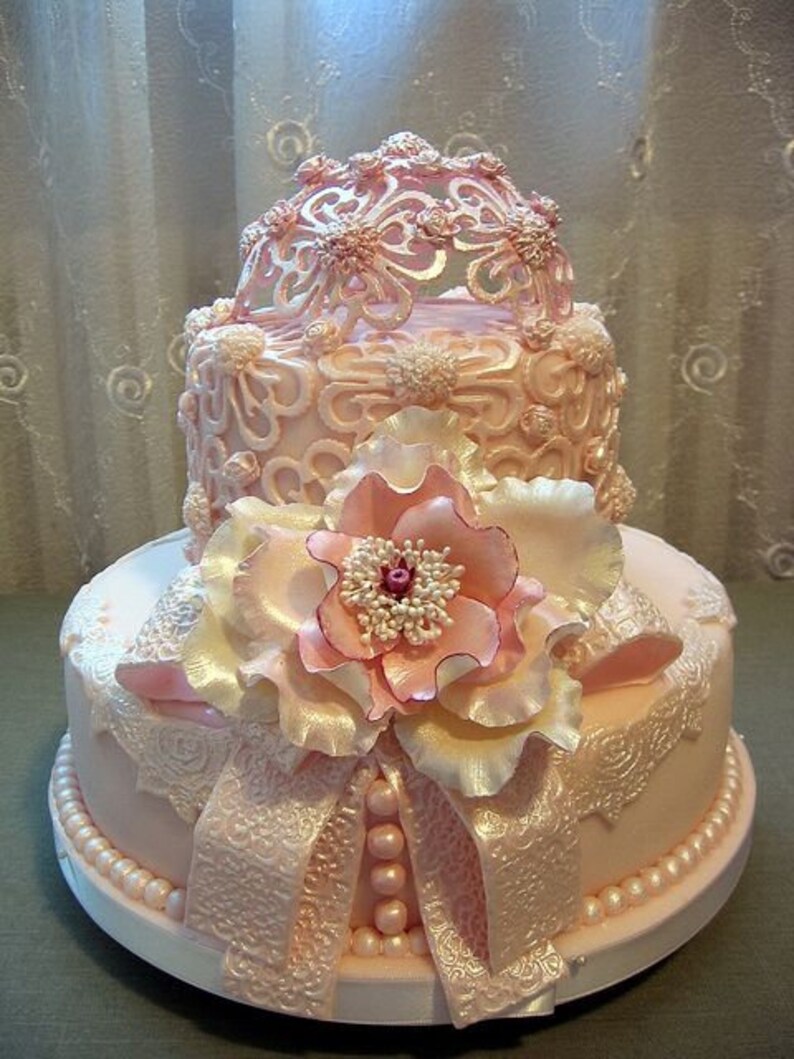 Peut inclure: Un g&acirc;teau de mariage &agrave; deux &eacute;tages de couleur rose, d&eacute;cor&eacute; de dentelle et de perles. Le niveau sup&eacute;rieur pr&eacute;sente un d&ocirc;me ajour&eacute; d&eacute;licat, tandis que le niveau inf&eacute;rieur est orn&eacute; d'une grande fleur rose.