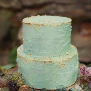 K&ouml;nnte beinhalten: Zweist&ouml;ckiger Kuchen mit hellblauem Buttercreme-Frosting und goldenen Blatt-Akzenten. Der Kuchen steht auf einem Baumstumpf, umgeben von kleinen Steinen.