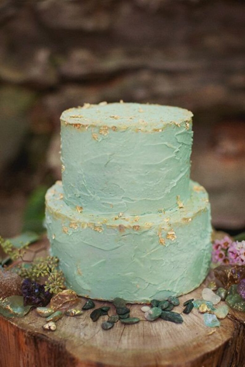K&ouml;nnte beinhalten: Zweist&ouml;ckiger Kuchen mit hellblauem Buttercreme-Frosting und goldenen Blatt-Akzenten. Der Kuchen steht auf einem Baumstumpf, umgeben von kleinen Steinen.
