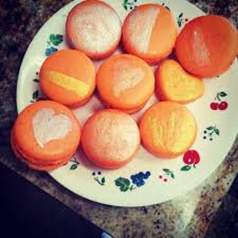 Peut inclure: Une assiette de neuf macarons orange avec diff&eacute;rents motifs dor&eacute;s et argent&eacute;s. Les macarons sont dispos&eacute;s en cercle sur une assiette blanche &agrave; motifs floraux.