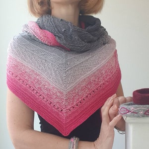 May include: A grey, pink and white triangular shawl with a lacy pattern. The shawl is being worn by a person with short blonde hair. The person is wearing a black top and a bracelet.
