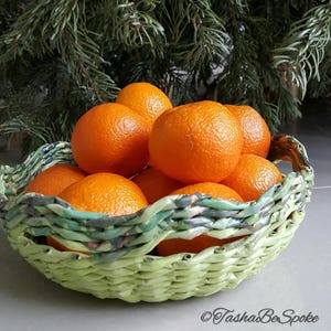 Puede incluir: Una cesta tejida verde claro llena de mandarinas de color naranja brillante. La cesta tiene un dise&ntilde;o tejido. Un &aacute;rbol de hoja perenne verde oscuro forma el fondo. Las mandarinas tienen una superficie texturizada.