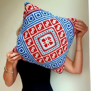 Op de afbeelding: Een vierkant gehaakt kussen met een geometrisch ontwerp in rood, wit en blauw. Het ontwerp heeft een centraal stervormig patroon, omgeven door concentrische vierkanten en rechthoeken. Het kussen wordt door een persoon vastgehouden.