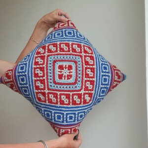 Op de afbeelding: Een vierkant, gehaakt kussen met een geometrisch ontwerp in rood, blauw en wit. Het kussen heeft een centraal vierkant met een bloemachtig patroon, omgeven door herhalende vierkante en rechthoekige vormen. Het kussen wordt door twee handen omhoog gehouden.