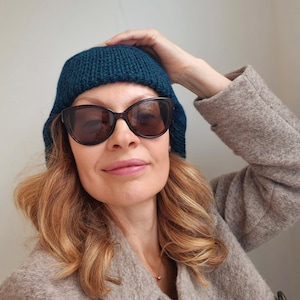 K&ouml;nnte beinhalten: Eine Frau tr&auml;gt eine tealblaue Strickm&uuml;tze und eine braune Sonnenbrille. Sie tr&auml;gt einen hellbraunen Mantel.
