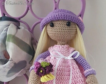 May include: A crocheted doll with blonde hair, wearing a purple hat, a pink dress with a white lace trim, and grey and purple boots. The doll is holding a bouquet of purple, green, and yellow flowers.