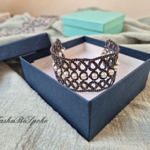 Puede incluir: Un delicado brazalete de encaje negro con detalles de perlas blancas, presentado en una caja de regalo azul marino. El brazalete est&aacute; hecho con intrincados patrones florales y es una hermosa pieza de joyer&iacute;a.