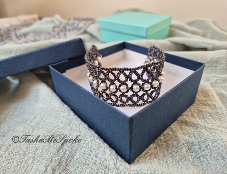Puede incluir: Un delicado brazalete de encaje negro con detalles de perlas blancas, presentado en una caja de regalo azul marino. El brazalete est&aacute; hecho con intrincados patrones florales y es una hermosa pieza de joyer&iacute;a.