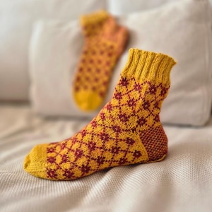 May include: A pair of knitted socks in a warm color palette of yellow and burgundy. The socks feature a geometric diamond pattern and ribbed cuffs. The second sock is slightly out of focus in the background, resting on a white surface.