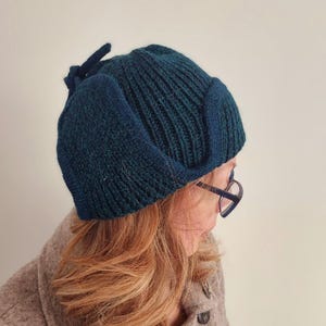 K&ouml;nnte beinhalten: Eine dunkelblaue und gr&uuml;ne Strickm&uuml;tze mit einer einzigartigen Form und einer kleinen Schleife oben. Die M&uuml;tze ist aus einem weichen, strukturierten Garn gefertigt.