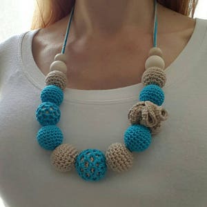Op de afbeelding: Een handgemaakte ketting met gehaakte en houten kralen. De ketting heeft afwisselend lichtblauwe en beige gehaakte kralen, met natuurlijke houten kralen. De ketting is geregen op een blauw-wit koord.