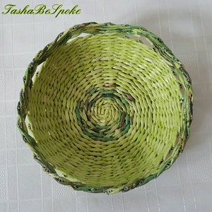 Puede incluir: Una cesta redonda hecha a mano, tejida con papel, con tonos verdes y un dise&ntilde;o en espiral en el centro. El borde de la cesta est&aacute; detallado con un ribete verde oscuro. El texto "TashaBeSpoke" es visible en la esquina superior izquierda.