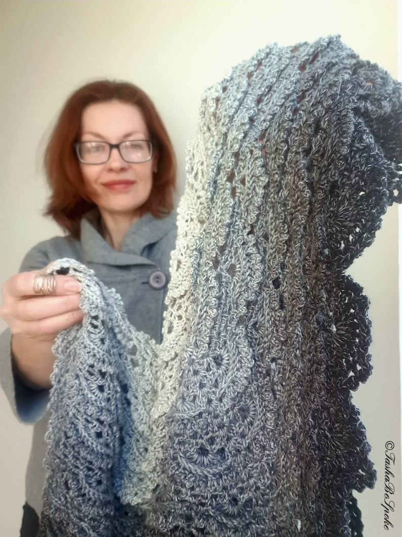 May include: A grey and white crocheted shawl with a scalloped edge. The shawl is made with a gradient yarn that transitions from light grey to dark grey.