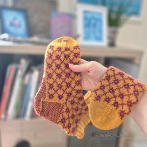 May include: A pair of hand-knitted socks in mustard yellow and burgundy. The socks feature a diamond pattern and are held up, showcasing the detailed craftsmanship. The socks are a cozy accessory.