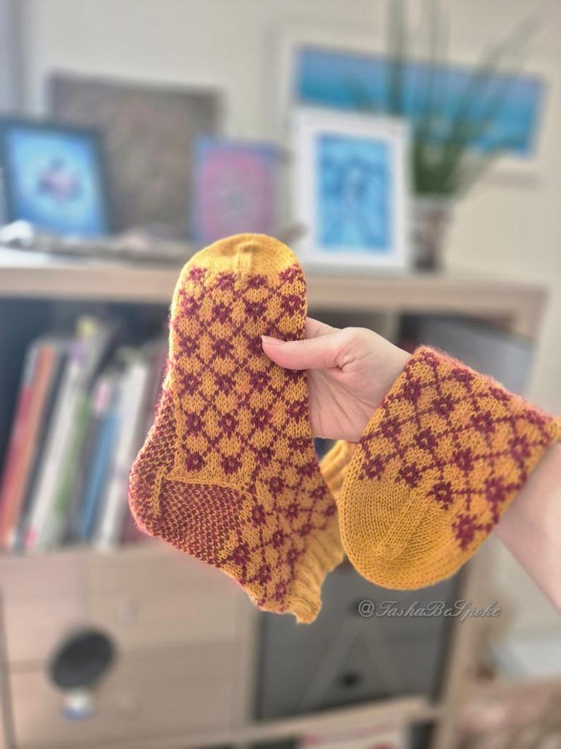 May include: A pair of hand-knitted socks in mustard yellow and burgundy. The socks feature a diamond pattern and are held up, showcasing the detailed craftsmanship. The socks are a cozy accessory.