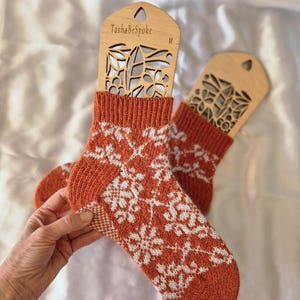 May include: A pair of orange and white patterned socks displayed on wooden sock forms. The socks feature a repeating white floral design on an orange background. The sock forms are labeled "TashaBeSpoke" and "M".