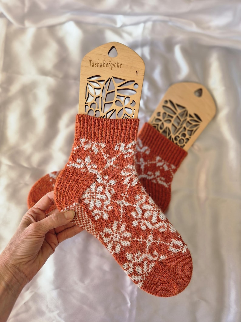 May include: A pair of orange and white patterned socks displayed on wooden sock forms. The socks feature a repeating white floral design on an orange background. The sock forms are labeled "TashaBeSpoke" and "M".