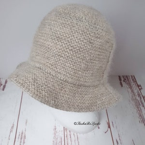 May include: A light beige knitted bucket hat with a slightly flared brim. The hat is made of a soft, textured yarn and has a rounded crown. The hat is displayed on a white mannequin head against a light wood background.