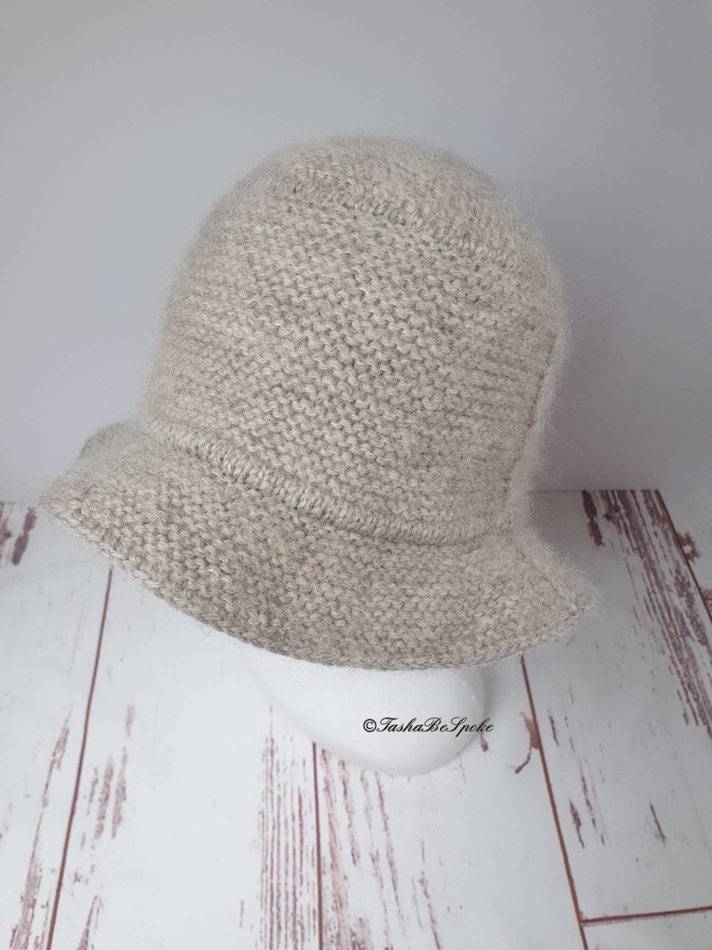 May include: A light beige knitted bucket hat with a slightly flared brim. The hat is made of a soft, textured yarn and has a rounded crown. The hat is displayed on a white mannequin head against a light wood background.