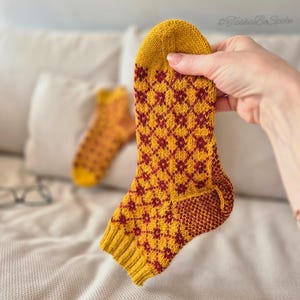 May include: A hand holds a yellow and burgundy patterned sock. The sock features a diamond pattern in burgundy against a yellow background. Another sock is visible in the background. The socks appear to be handmade and knitted.