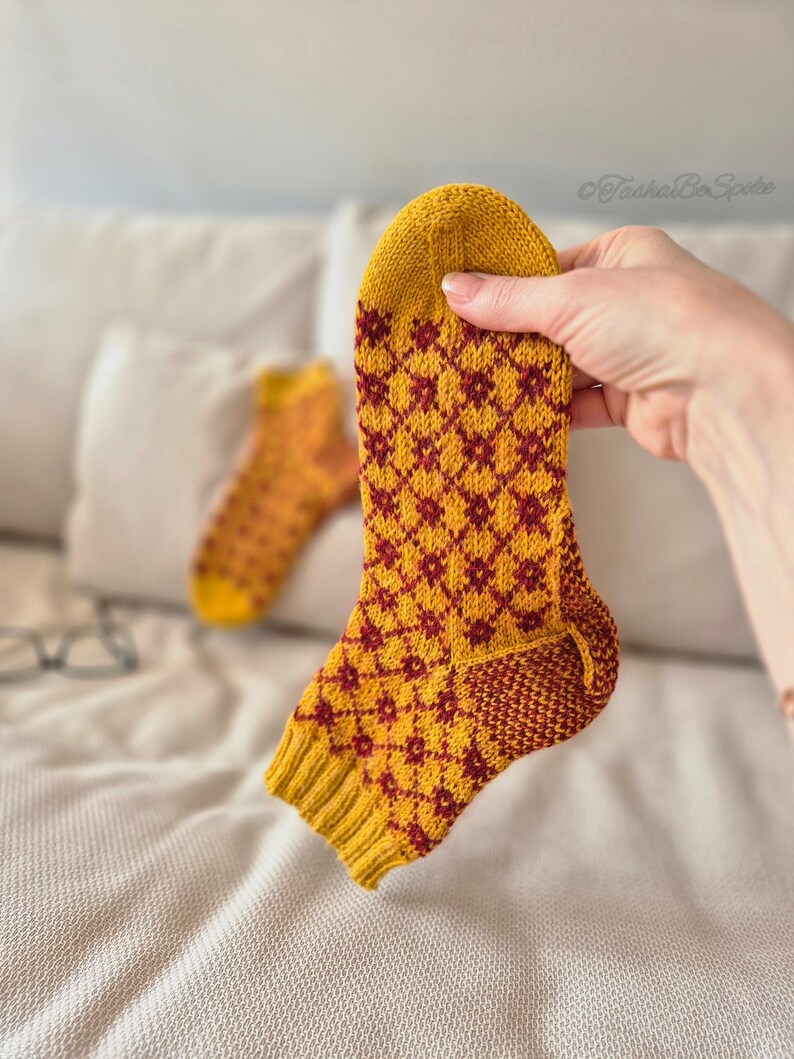 May include: A hand holds a yellow and burgundy patterned sock. The sock features a diamond pattern in burgundy against a yellow background. Another sock is visible in the background. The socks appear to be handmade and knitted.