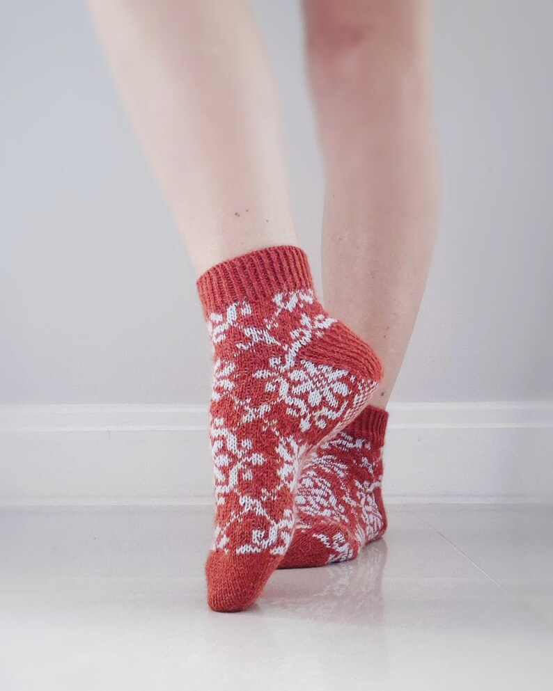 May include: A pair of red and white patterned socks. The socks feature a repeating white snowflake design on a red background. The socks are knit and appear to be made of a soft, warm material. The socks are worn on a person's feet.