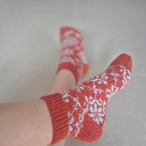 May include: A pair of coral-colored socks with a white snowflake pattern. The socks are knit and have ribbed cuffs. The image shows the socks on a person's feet.