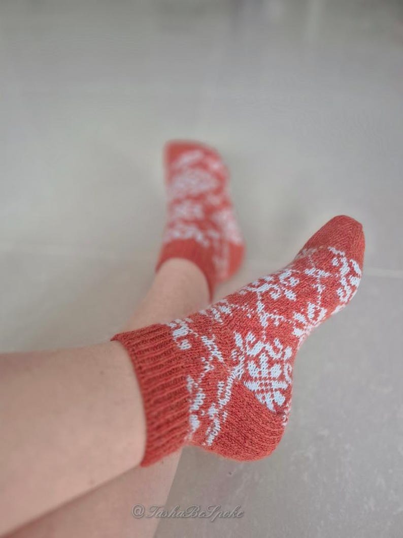 May include: A pair of coral-colored socks with a white snowflake pattern. The socks are knit and have ribbed cuffs. The image shows the socks on a person's feet.
